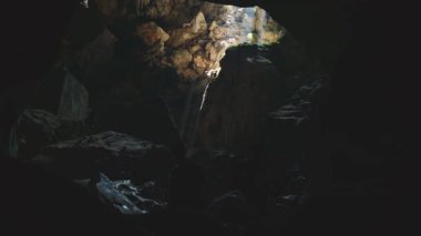 God rays illuminate majestic Laos waterfall flowing through a dark, mysterious, ancient underground cave entrance.