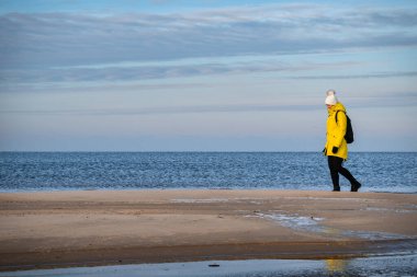 Sarı kışlık ceketli ve siyah sırt çantalı bir kadın Letonya, Carnikava yakınlarında Baltık Denizi kıyısında yürüyor.