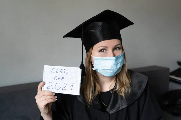 Graduate girl in medical mask at coronavirus covid-19 period. Woman with master degree in black graduation gown and cap. Distance learning online. Study at home. Graduation college.