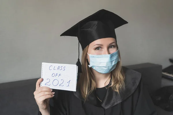 Graduate girl in medical mask at coronavirus covid-19 period. Woman with master degree in black graduation gown and cap. Distance learning online. Study at home. Graduation college.