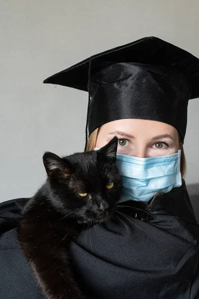 Graduation of a woman with face mask dressed in a black gown and holding black cat