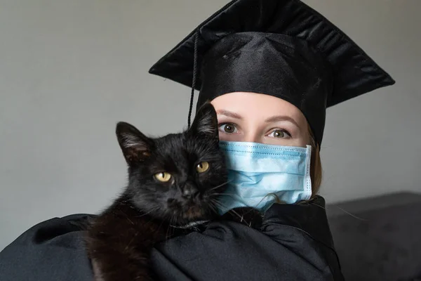 Siyah bir elbise giymiş ve elinde siyah bir kedi tutan yüz maskeli bir kadının mezuniyeti.