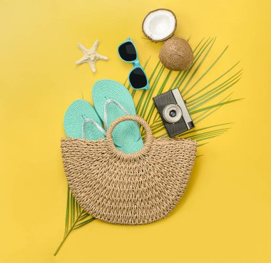 Bir plaj çantası, parmak arası terlik, kamera, palmiye yaprağı, güneş gözlüğü, hindistan cevizi ve deniz yıldızı karışımı sarı arka planda. yaz konsepti