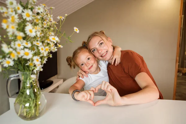 Mom and daughter showing heart symbol with their hands. Spending time with family at home concept. Bouquet of chamomiles in front of the girls.