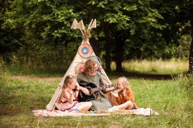 Annem ormanda çadırın yanında oturan küçük kızına gitar çalar. Aile yaz tatillerinde eğleniyor, müzik festivalinde. Küçük kızlar ellerini çırpar.