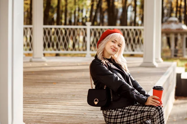 Trendige Frau in Lederjacke, karierter Hose, roter Baskenmütze mit roter Kaffeetasse. — Stockfoto