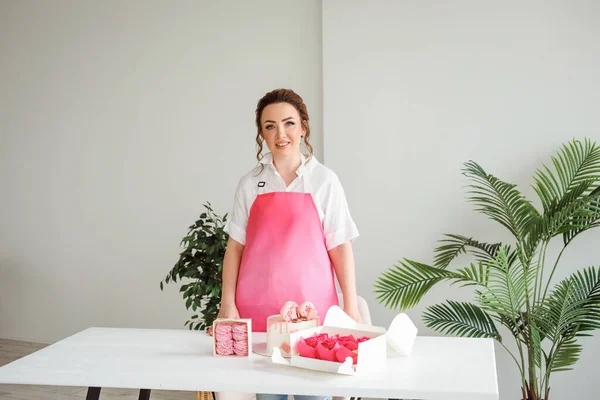蛋糕店的厨师穿着粉红围裙 坐在白桌上美味的甜点旁边 — 图库照片