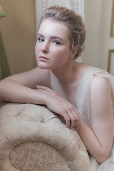 Mild porträtt av en ung vacker flicka i en ljus chiffong klänning med ljus sminkning och frisyr sitter på soffan foton i milda toner i stil med konst — Stockfoto
