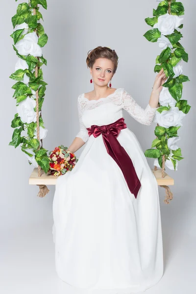 Bela noiva adorável está sentado em um balanço com um belo buquê de flores coloridas em um vestido branco com penteado à noite — Fotografia de Stock