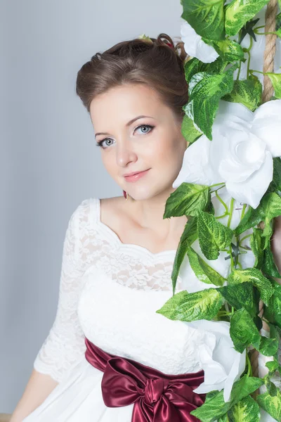 Beautiful lovely bride is sitting on a swing with a beautiful bouquet of colorful flowers in a white dress with evening hairstyle — Stock Photo, Image