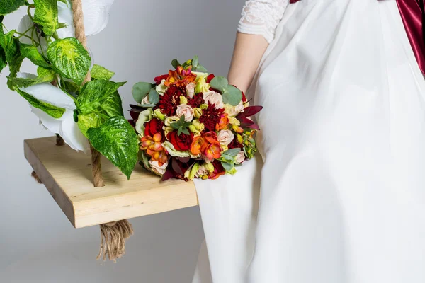 Hermosa novia encantadora está sentado en un columpio con un hermoso ramo de flores de colores en un vestido blanco con peinado de noche — Foto de Stock