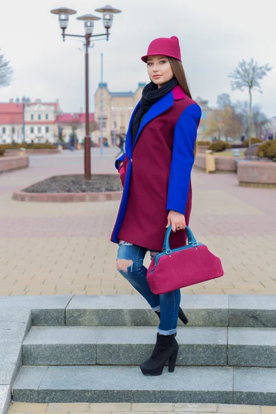 Beautiful happy cute girl with long hair in a hat and bright colored coat with a bag walks through the city in a bright sunny day — Stock Photo, Image