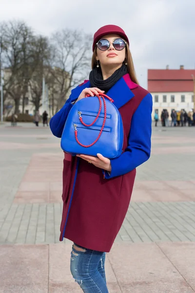 Vackra glad söt tjej med långt hår i en hatt och ljusa färgade kappa med en påse går genom staden i en solig dag — Stockfoto