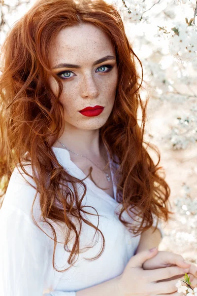 Niedliches Porträt eines schönen rothaarigen Mädchens mit Make-up und rotem Lippenstift in einem weißen Hemd im Garten zwischen blühenden Bäumen an einem sonnigen, warmen Abend — Stockfoto