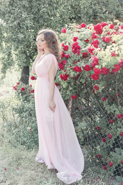 Hermosa chica sexy en un vestido ligero con maquillaje delicado y cabello en un jardín de flores con rosas en luchas la luz del sol al atardecer. Estilo foto fane arte — Foto de Stock