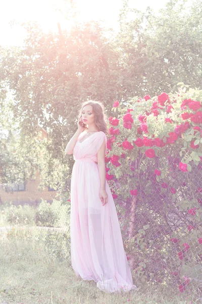 Bella ragazza sexy in un abito leggero con delicato make-up e capelli in un giardino fiorito con rose alla luce del sole luchas al tramonto. Foto in stile fane art — Foto Stock