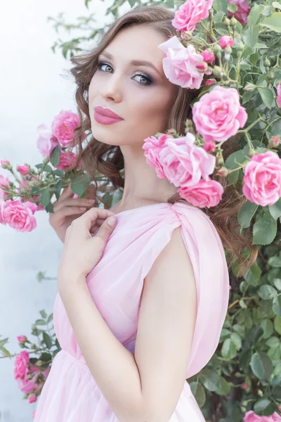 Belle fille sexy dans une robe rose debout dans les roses du jardin dans une journée d'été ensoleillée et lumineuse avec un maquillage doux et de grandes lèvres bouffies lumineuses — Photo