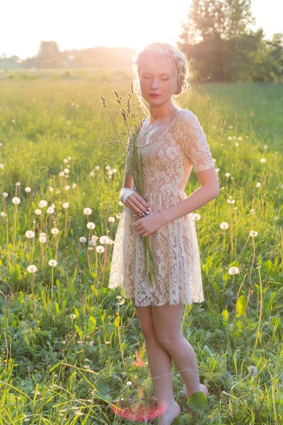 Schöne junge süße Mädchen in einem weißen Kleid mit Haaren auf dem Kopf schräg geht in einem Feld bei Sonnenuntergang — Stockfoto