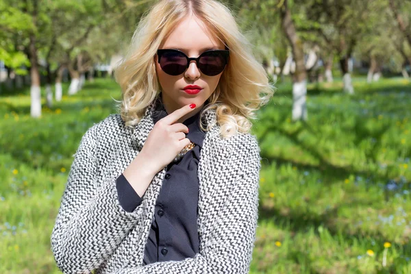 Mooi sexy blond meisje met rode lippen in zonnebril wandelen in de tuin van een zonnige dag — Stockfoto