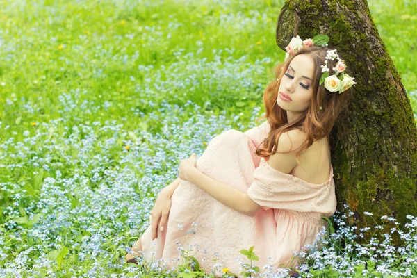 Krásná sexy dívka s červenými vlasy s květinami ve vlasech, sedí u stromu v růžových šatech na louce s modrými květy — Stock fotografie