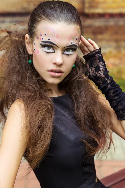 Mooi meisje op de achtergrond van de bladeren in de herfst dag op straat met fantasie make-up in een zwarte jurk met grote sexy lippen — Stockfoto