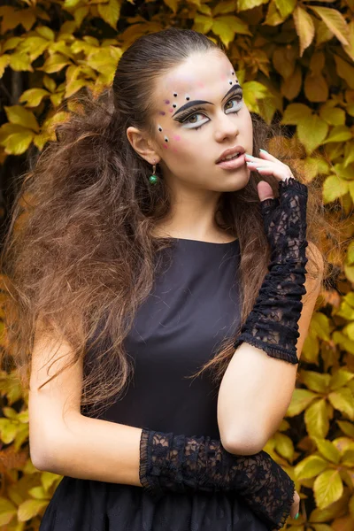 Mooi meisje op de achtergrond van de bladeren in de herfst dag op straat met fantasie make-up in een zwarte jurk met grote sexy lippen — Stockfoto