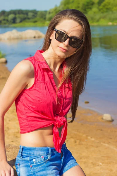 Сексуальная красивая девушка с длинными темными волосами в солнцезащитных очках, сидящая в джинсовых шортах на пляже у воды в солнечный день — стоковое фото