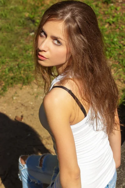 Beautiful sexy girl with long hair in a white t-shirt and jeans sitting in the woods on a Sunny day — Stock Photo, Image