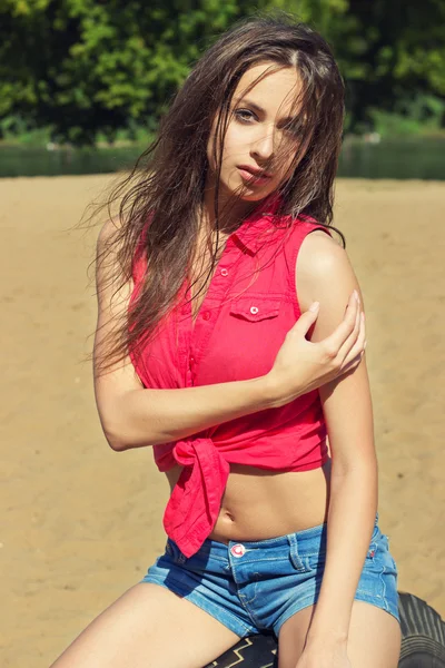 Sexy piękna dziewczyna z długimi ciemnymi włosami siedzi w dżinsowych spodenkach na plaży w słoneczny dzień — Zdjęcie stockowe