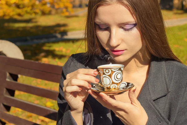 Schöne Frau mit einer Tasse Tee im schönen Make-up sitzt im Park auf einer Bank und trinkt heißen Tee im Herbst sonnigen Tag — Stockfoto