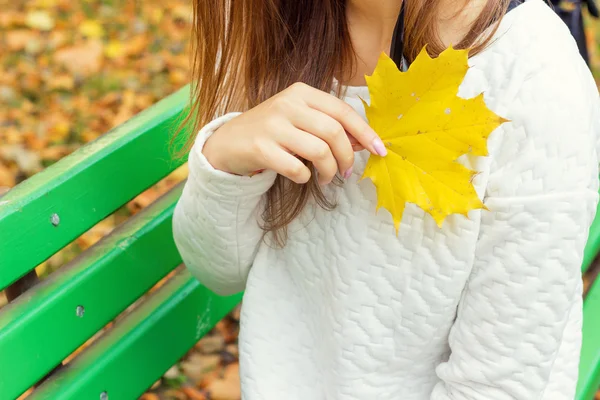 Schönes Mädchen in weißer Jacke und schwarzer Hose mit gelben Blättern in den Händen, die auf der Bank im Herbstpark sitzen — Stockfoto