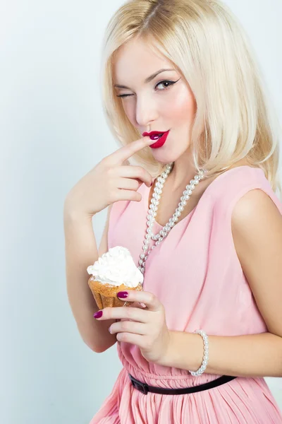 Schöne sexy elegante blonde Frau mit hellem Make-up in rosa Kleid im Studio auf weißem Hintergrund sitzt mit einem Cupcake in den Händen und leckt die süße Sahne von seinem Finger zwinkert Retro-Stil — Stockfoto