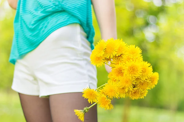 Hermosa chica en un día soleado de verano caminando en el jardín y mantiene dientes de león amarillos en las manos — Foto de Stock