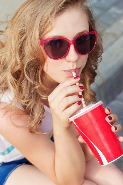 Hermosa joven en gafas de sol sentado en la calle y beber una coca con una taza de papel del tubo —  Fotos de Stock