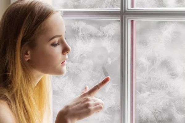 Hermosa joven sentada junto a la ventana el día de invierno y dibuja el sol en la ventana congelada — Foto de Stock