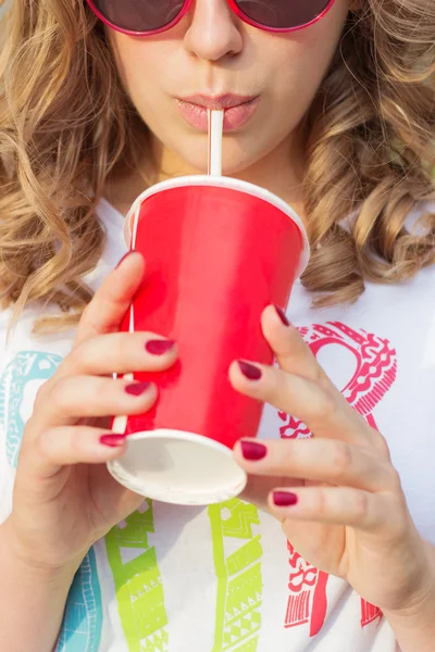 Beautiful young girl in sunglasses in the summer warm day drinking coke through a straw with red glass — Stock Photo, Image