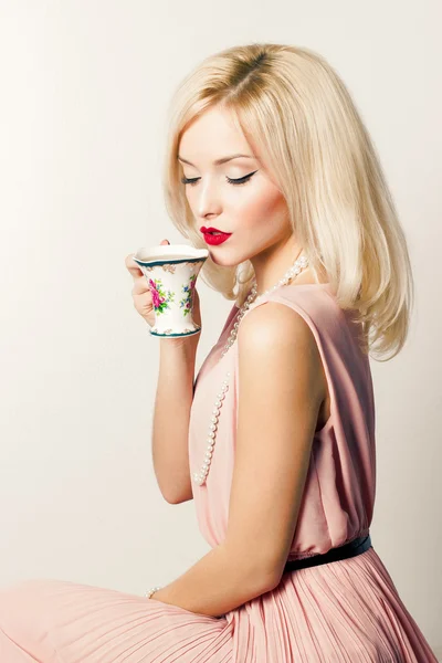 Hermosa sonrisa feliz chica elegante sexy con lápiz labial rojo en un vestido rosa en estilo retro bebe café de té de una taza pequeña —  Fotos de Stock