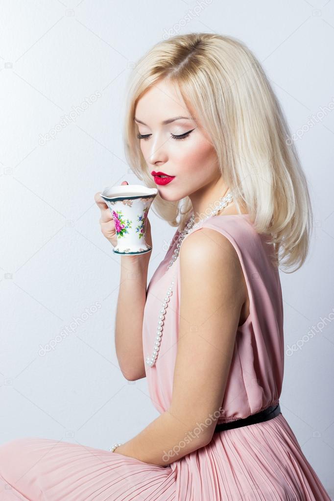 Beautiful smiling happy sexy elegant girl with red lipstick in a pink dress in retro style drinks tea coffee from a small mugs