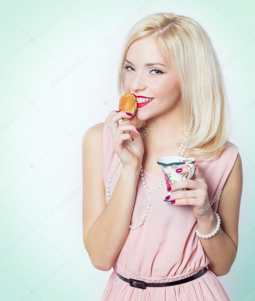 Beautiful sexy gorgeous blonde girl with bright makeup in pink dress in the Studio on a white background sitting with a small mug in his hands and winks and bite of croissant pastry, retro style