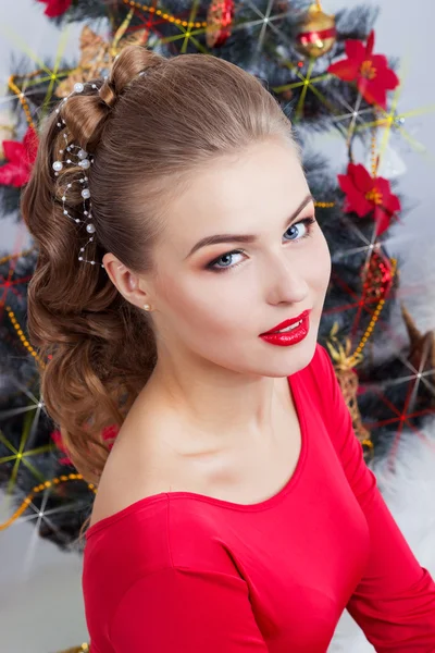 Hermosa sexy mujer joven sonriente feliz en vestido de noche con maquillaje brillante con lápiz labial rojo sentado cerca del árbol de Navidad en una noche de Navidad festiva —  Fotos de Stock