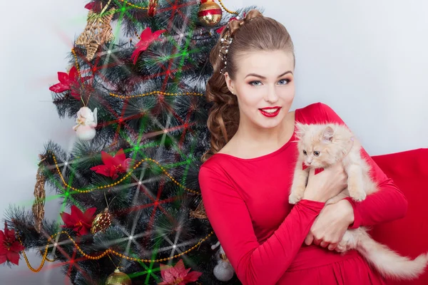 Vackra sexiga glada leende ung kvinna i aftonklänning med ljusa makeup med rött läppstift, sitter av granen med en liten kattunge i famnen i en festlig jul kväll — Stockfoto