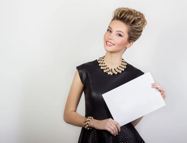 Retrato de una hermosa joven sexy feliz sonriendo en un vestido de noche negro con pelo y maquillaje con joyas caras con un signo blanco en sus manos en el estudio sobre fondo blanco —  Fotos de Stock