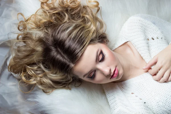 Beautiful smiling happy girl with bright makeup lies on the bed with fur in white sweater — Stock Photo, Image