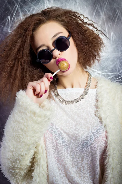 Divertido hermosa chica sexy en gafas y un abrigo blanco lame una barra de caramelo, maquillaje brillante, estudio de fotografía de moda — Foto de Stock
