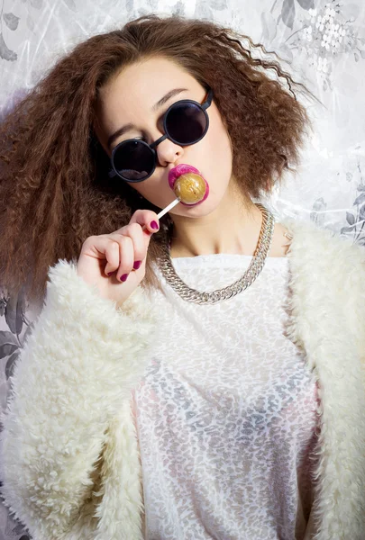Divertente bella ragazza sexy in occhiali e un cappotto bianco lecca una barretta di caramelle, trucco luminoso, fotografia di moda Studio — Foto Stock