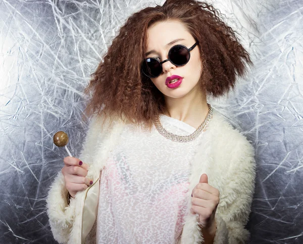 Beautiful girl with curly hair and bright lips in a white coat in the round sunglasses with a candy in his hands, Studio shot — Stock Photo, Image