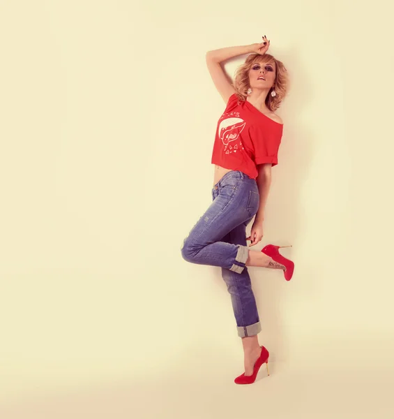 Hermosa chica sexy en jeans y una camiseta naranja estaba de pie cerca de una pared blanca, maquillaje brillante y peinados, estudio de fotos —  Fotos de Stock