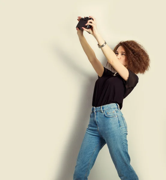 Beautiful young girl hipster takes photos, shoots selfe, taking pictures of himself on camera in jeans and a black t-shirt in the Studio — Stock Photo, Image
