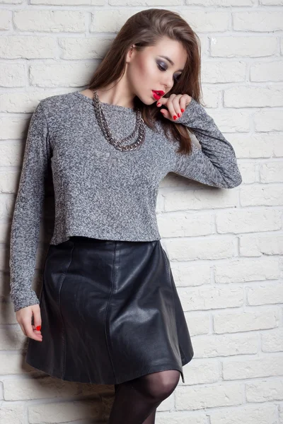 Beautiful young sexy girl with plump red lips with a red lacquer on the nails in a black leather skirt with a stylish haircut stands near brick wall — Stock Photo, Image
