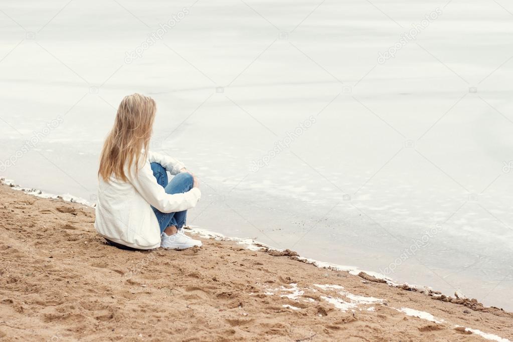 Beautiful young blonde girl in jeans and a white shirt sitting on the shore of the frozen cold of the lake near the forest in early spring